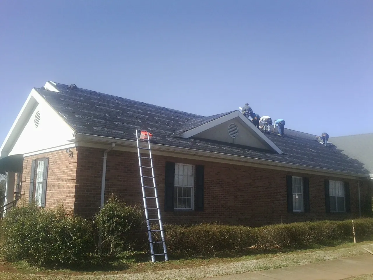 Crew tearing shingles off a brick ranch roof with a ladder set — Metal Roof Repair work by our crew in Cayce